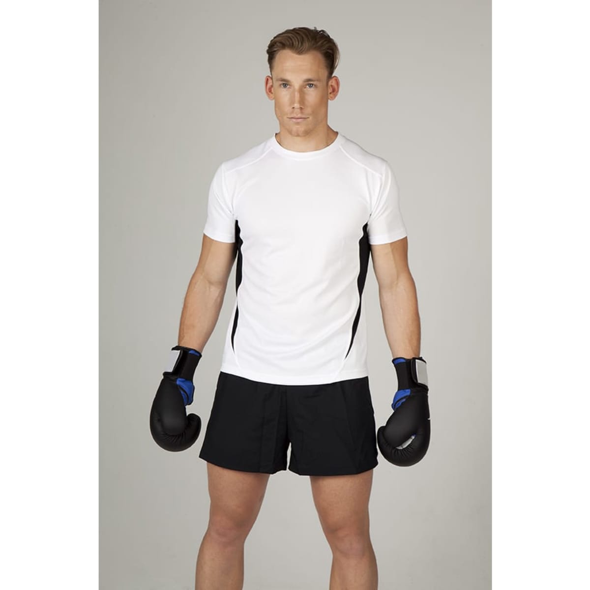 A man poses in a white and black short-sleeved T-shirt, wearing boxing gloves and black shorts.