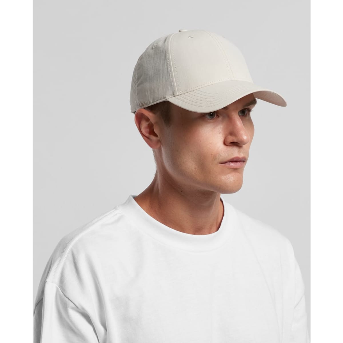 A beige nylon cap with a curved brim, worn by a model in a white t-shirt, featuring a logo on the front.