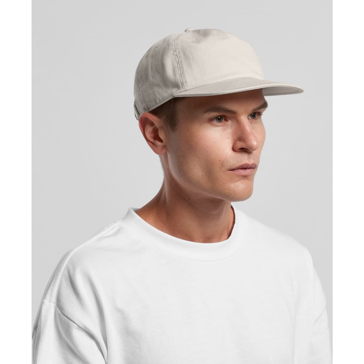 A light grey cap with a flat brim worn by a person in a white t-shirt, featuring a logo on the side.