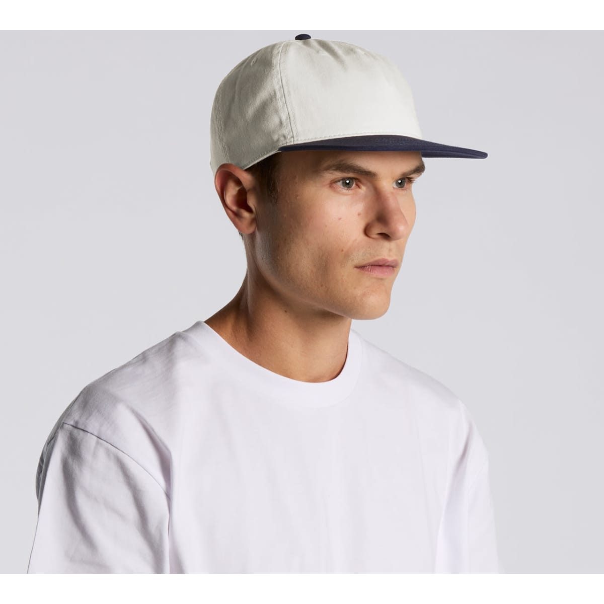 A cap featuring a two-tone design with a light-coloured crown and a navy peak, worn by a model.