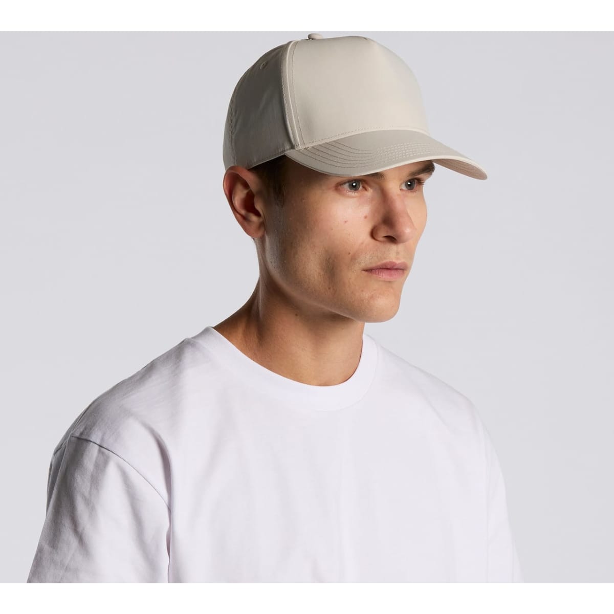 A beige nylon cap with a structured design worn by a person in a plain white T-shirt. It features a logo on the front.