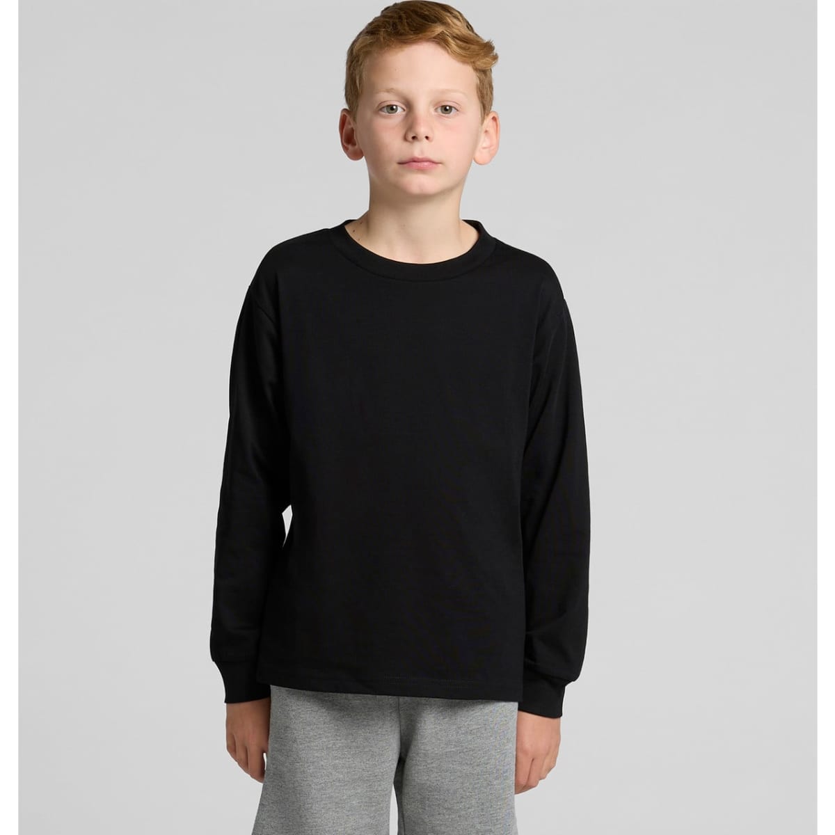 A black long-sleeve t-shirt for youth modelled by a child, paired with grey pants. The shirt features a logo.