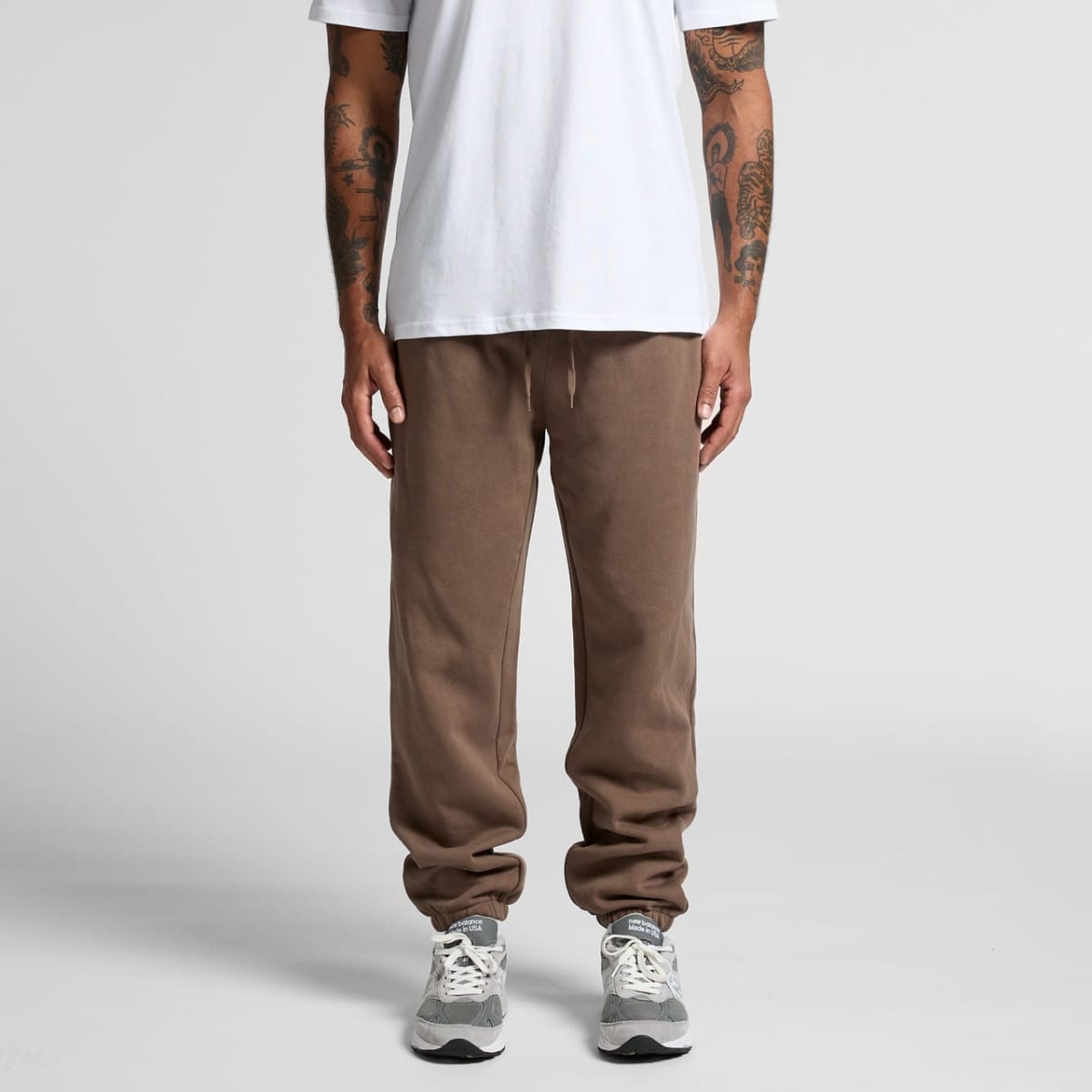 Brown track pants with a relaxed fit and an elastic waistband, paired with grey sneakers. The model wears a plain white t-shirt.