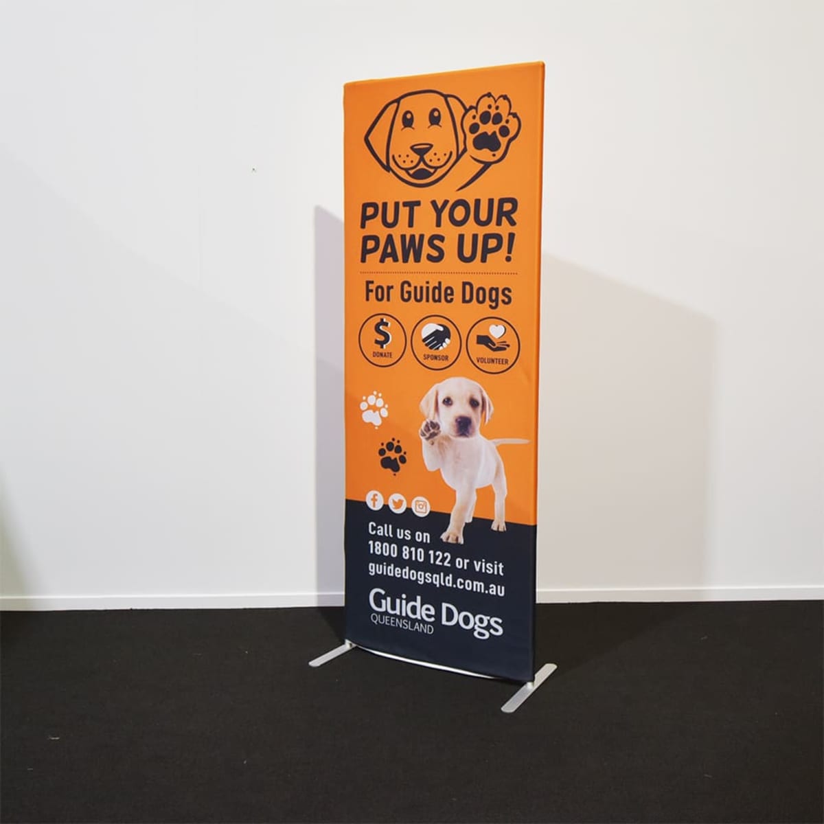 A promotional banner stand featuring an orange and black design, highlighting Guide Dogs Queensland with a logo.