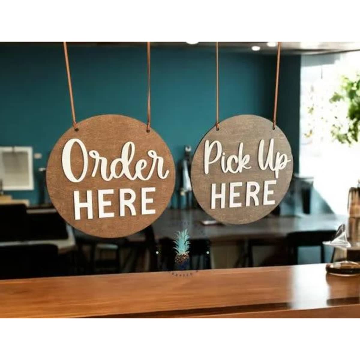 Two round wooden signs hang from a ceiling, one saying "Order Here" and the other "Pick Up Here," in white text.