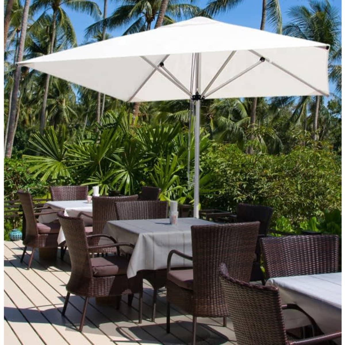 A white café umbrella with a sturdy frame stands over outdoor tables surrounded by lush greenery and palm trees.