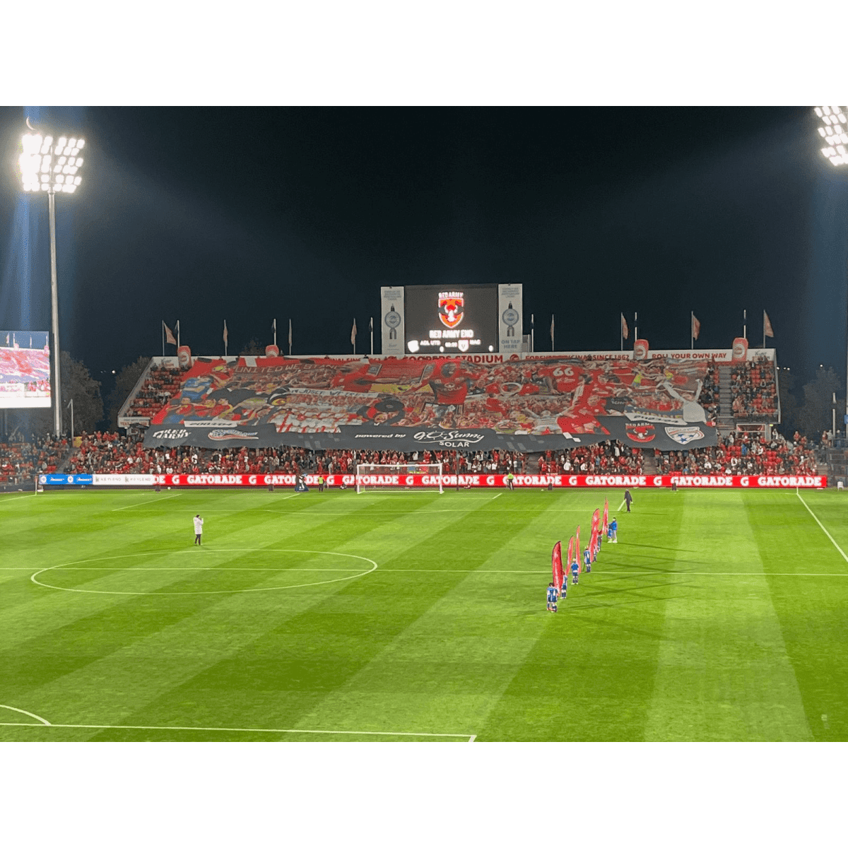 A large polyester TIFO banner in vibrant colours displayed at a stadium, with fans and a field in the foreground.