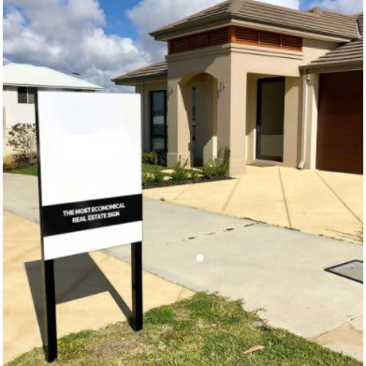 A flat metal sign on a black post stands outside a modern beige house on a paved driveway.
