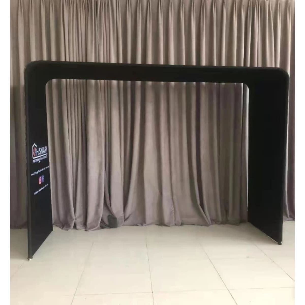 A black fabric arch frame with a tension design, set against a neutral curtain backdrop. Features a logo on one side.