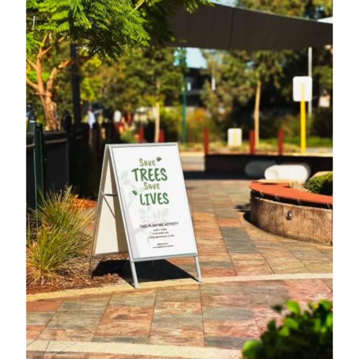 A-frame sign in white and silver displaying a message about saving trees, set in a landscaped outdoor area.