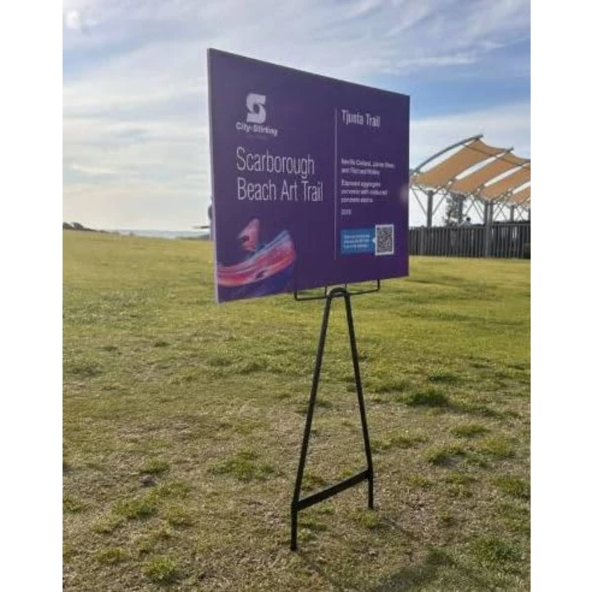 A promotional display featuring a purple sign with information about the Scarborough Beach Art Trail, on a black wire stand.