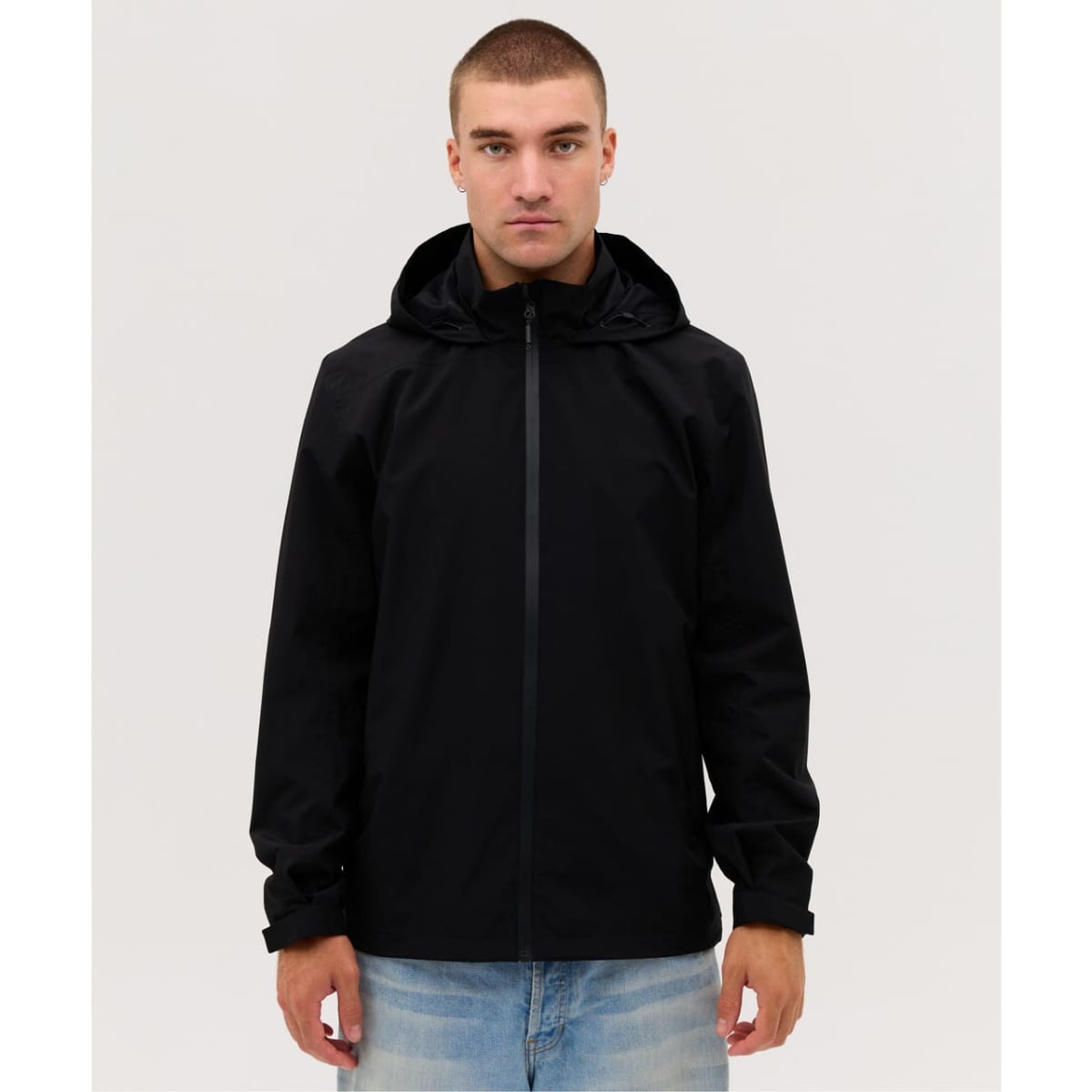 A black rain jacket with a hood, featuring a front zip and a sleek design. The model is wearing light jeans.