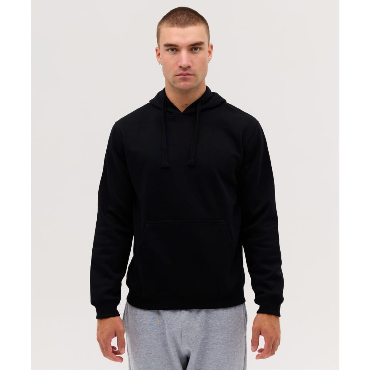 A black hooded jumper with a front pocket and drawstrings, worn by a model. The model is also wearing grey pants.