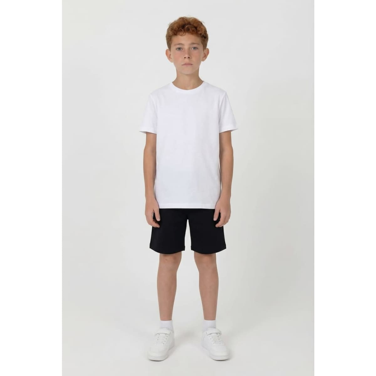 A boy wearing a white t-shirt and black sports shorts, standing on a plain background. The shorts have a logo.
