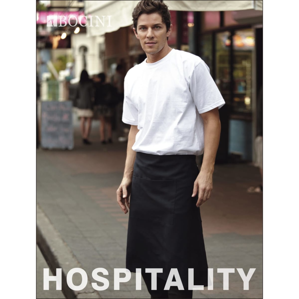 A man wears a black cotton drill three-quarter apron over a white T-shirt, standing in an outdoor setting. The apron features a pocket.