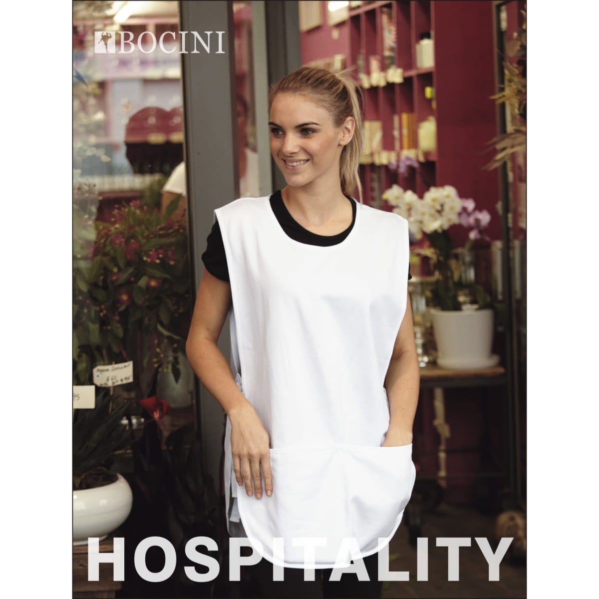 A white cotton drill popover apron with a pocket, displayed in a vibrant shop setting.