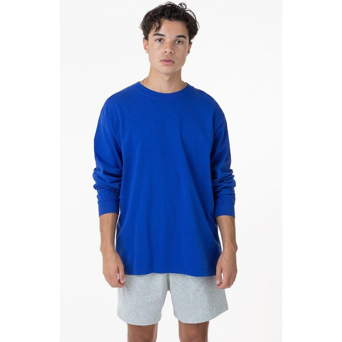 A long sleeve crewneck tee in bright blue, featuring a relaxed fit. The model wears grey shorts.