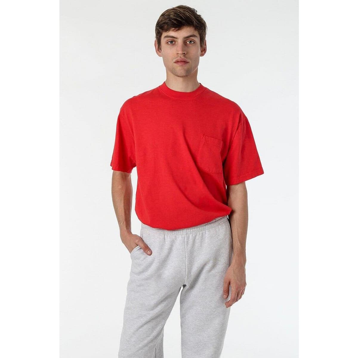 A red short sleeve T-shirt featuring a chest pocket, worn with grey sweatpants, with a logo visible on the shirt.