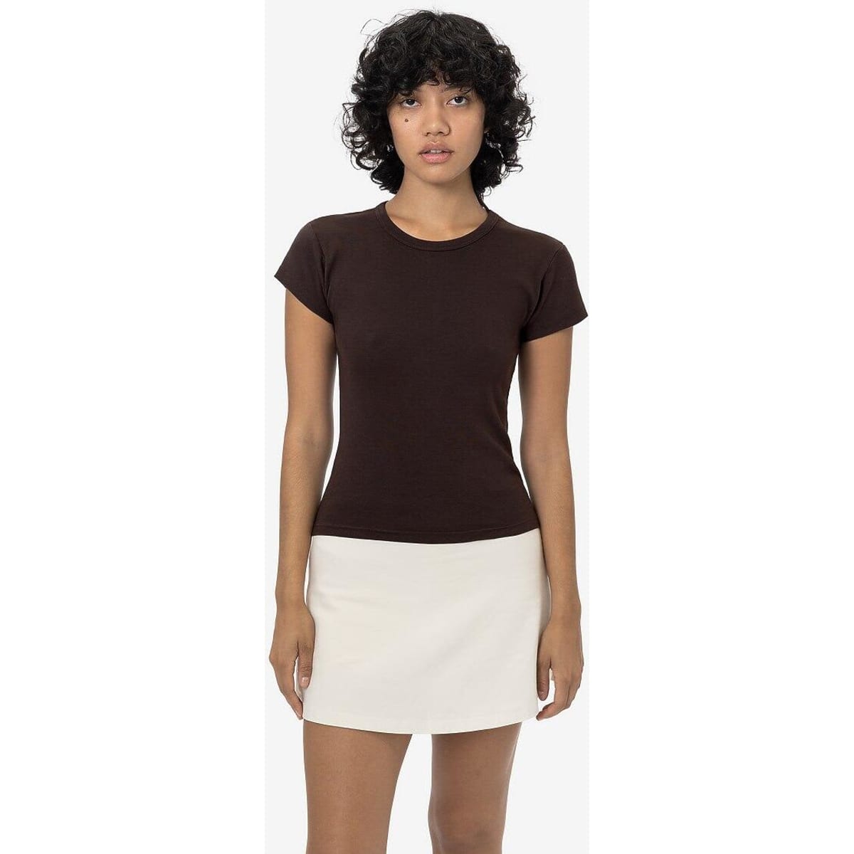 A brown cap sleeve baby tee displayed on a model, paired with a white skirt. The shirt has a subtle logo.