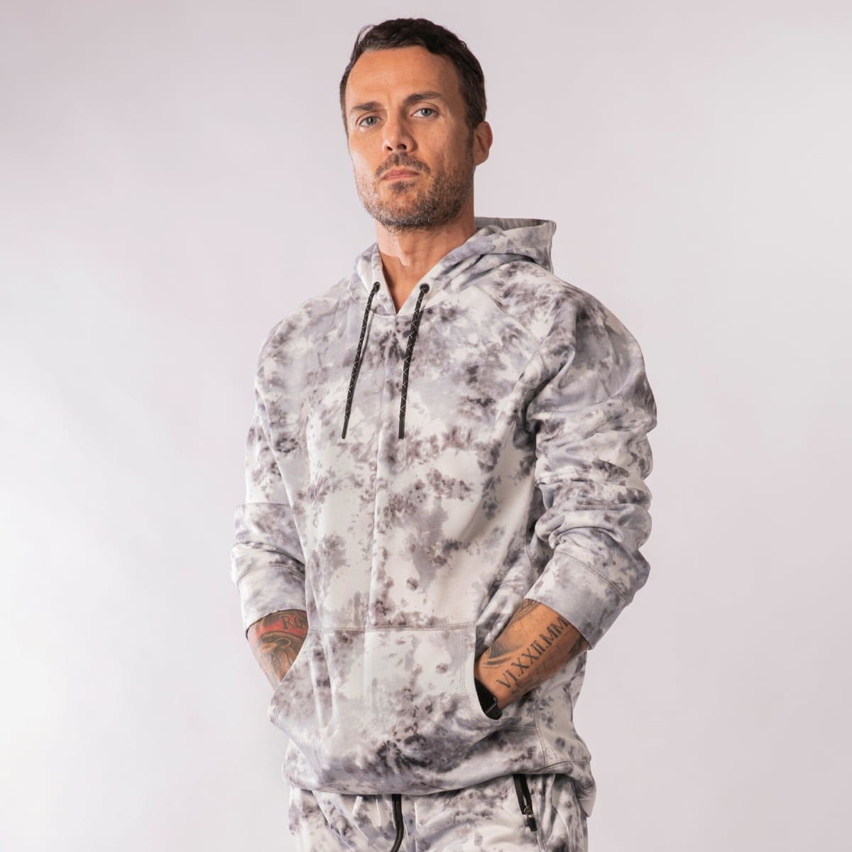 A tie-dye patterned pullover hoodie in grey tones, featuring a logo and a drawstring hood.