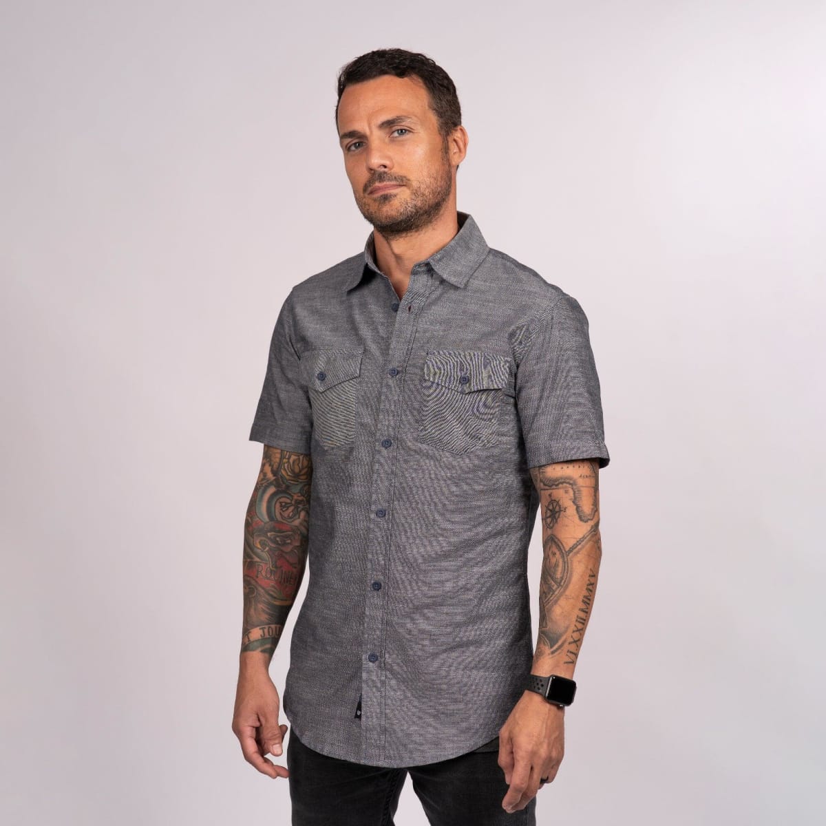 A man wearing a short sleeve chambray shirt in grey, featuring a pocket and a logo.
