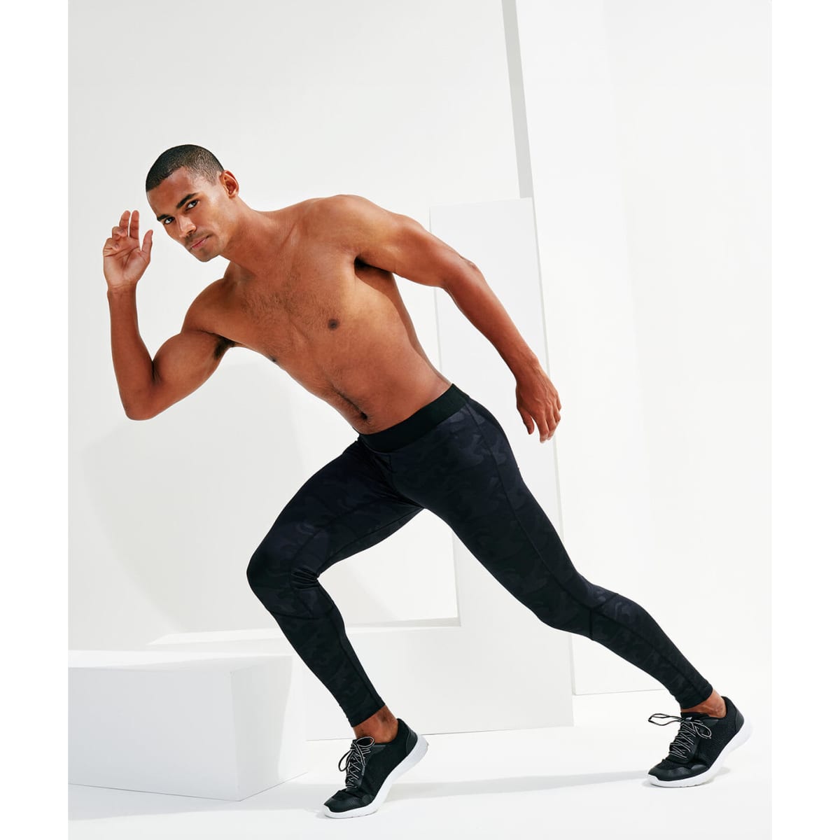 A man wearing black training leggings and black shoes, posed in an athletic stance against a bright background.
