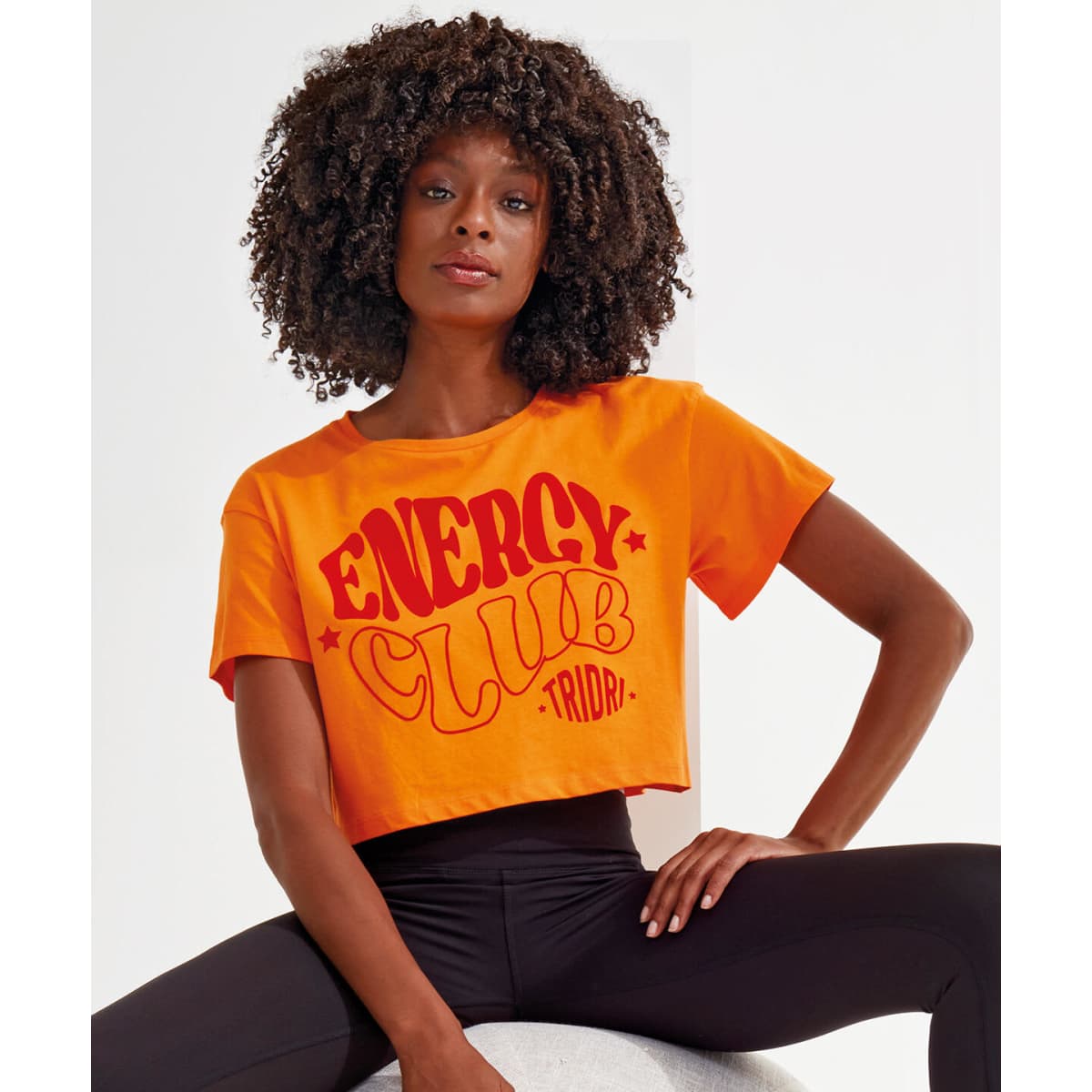 A model wearing an orange crop top with red text, featuring a logo, paired with black leggings.