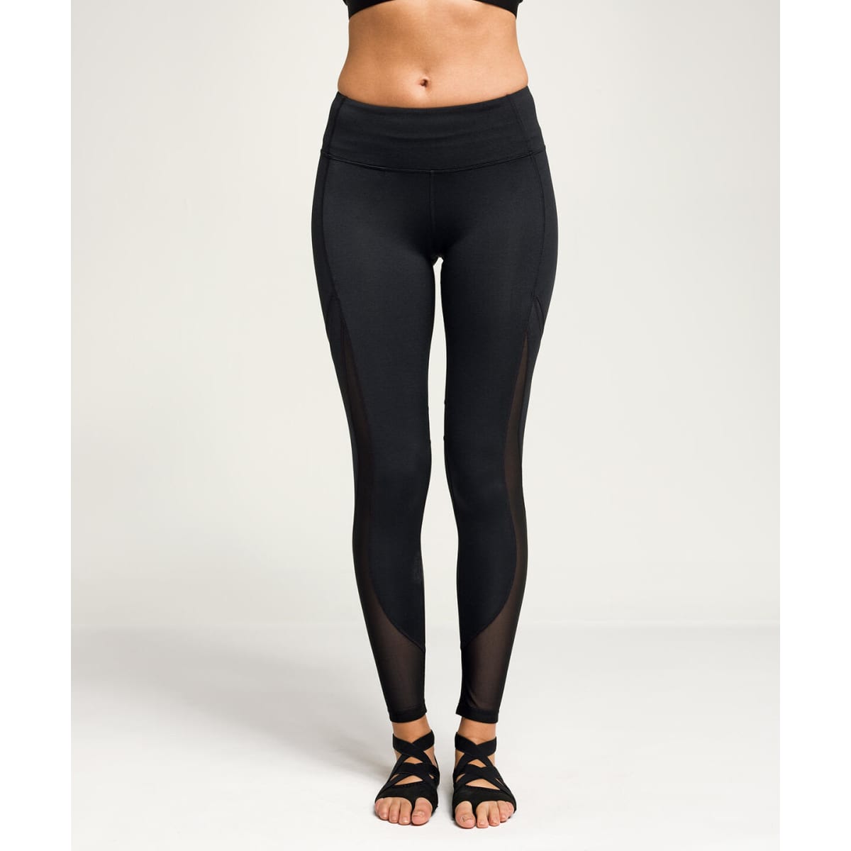 Full-length black leggings featuring mesh panels on the sides. The model is wearing sandals. Branding is present.
