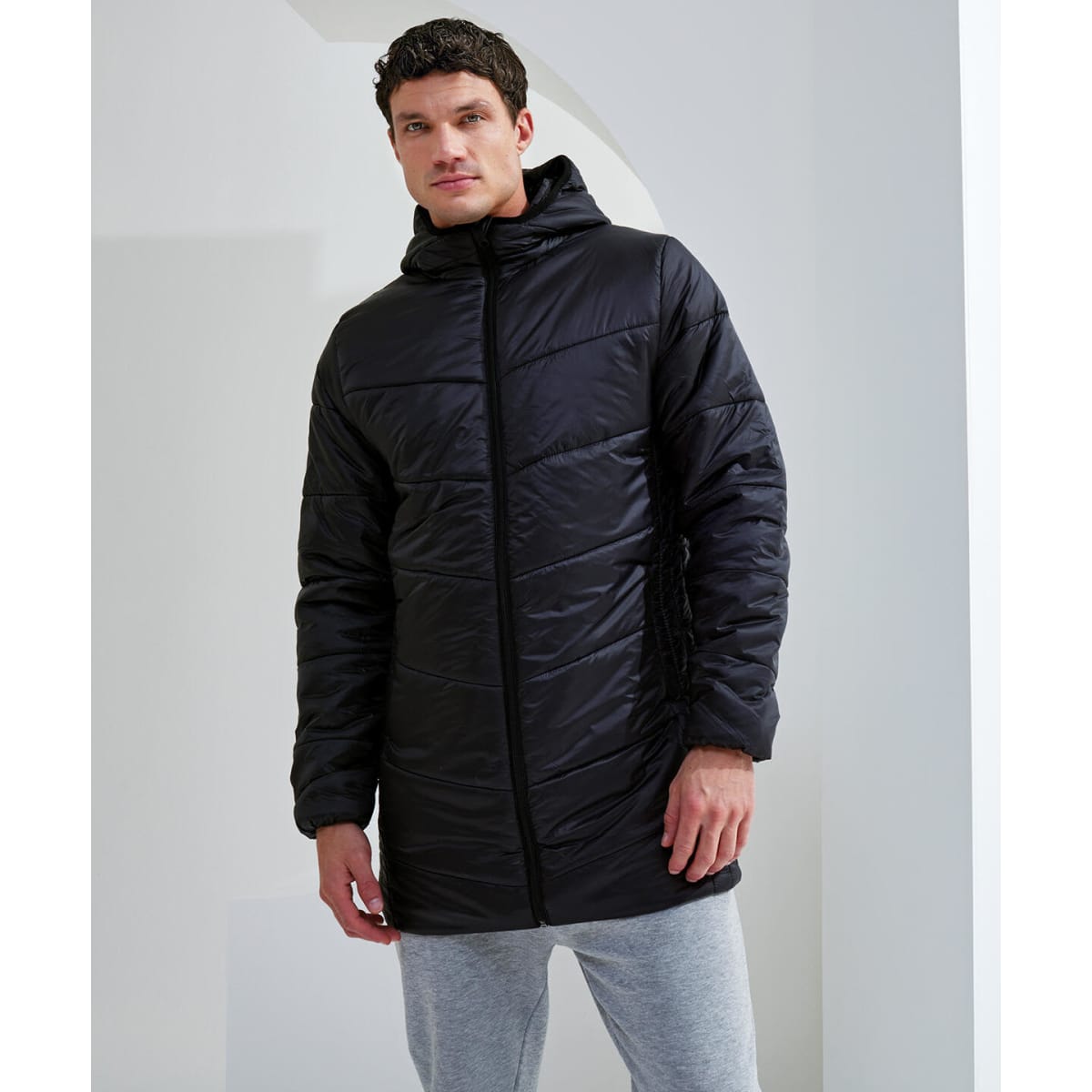A model wearing a black Microlight Longline Jacket with a hood, paired with grey pants, stands in an indoor setting.