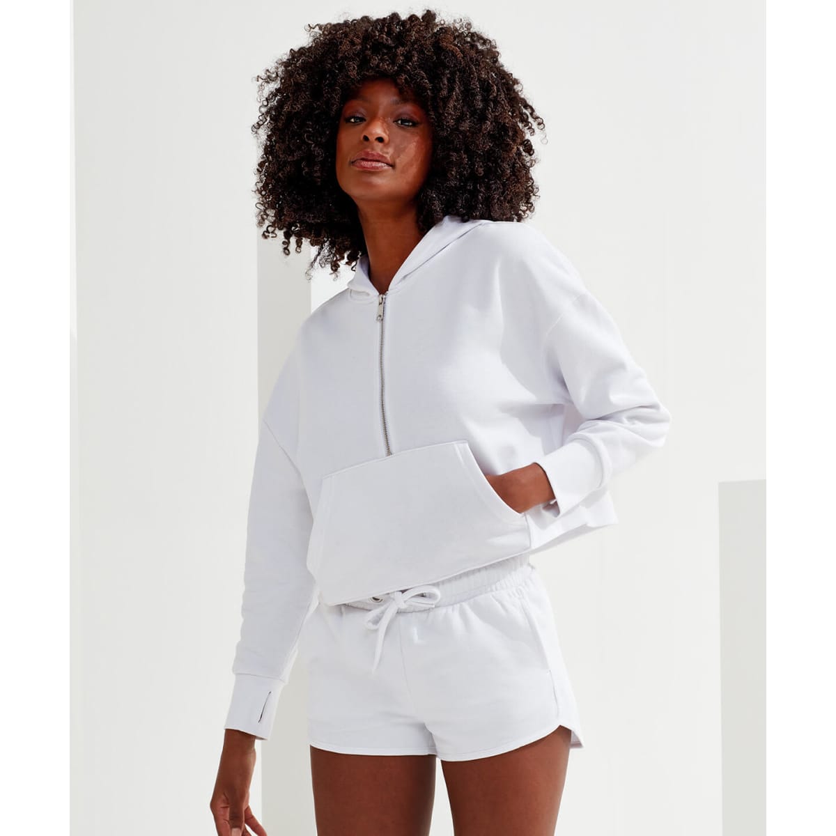 A white 1/2 zip hoodie featuring a front pocket, styled with matching shorts. The model has curly hair.