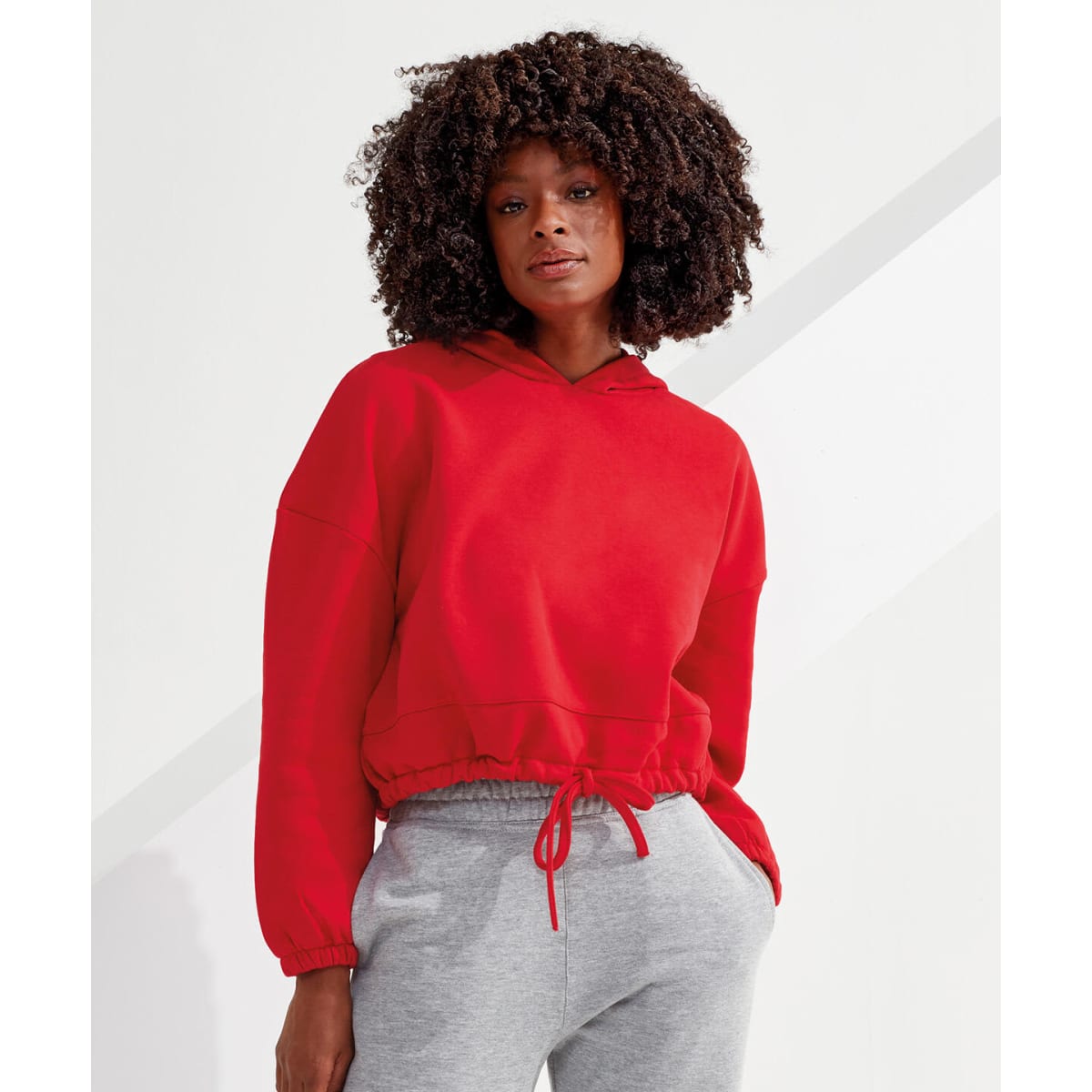 A red cropped oversize hoodie with a drawstring hood, paired with grey pants, featuring a logo on the fabric.