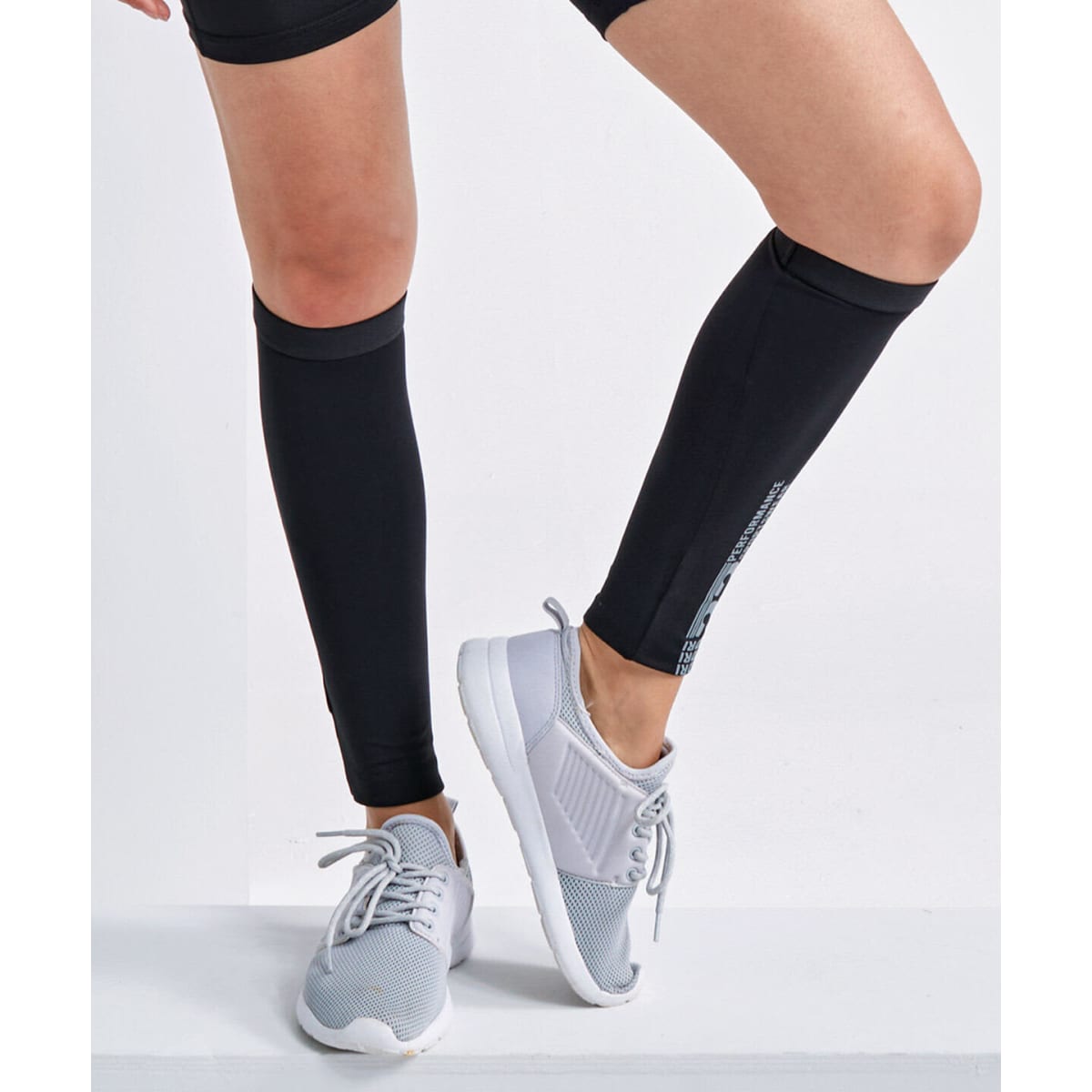 Black compression calf sleeves worn on legs, paired with light grey athletic shoes. The product features a logo.