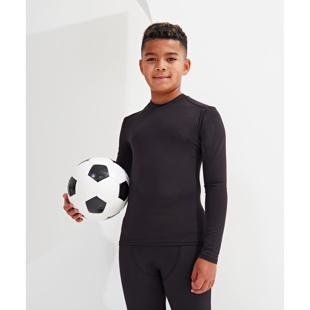 A boy stands holding a black and white soccer ball, wearing a fitted black performance baselayer with long sleeves.