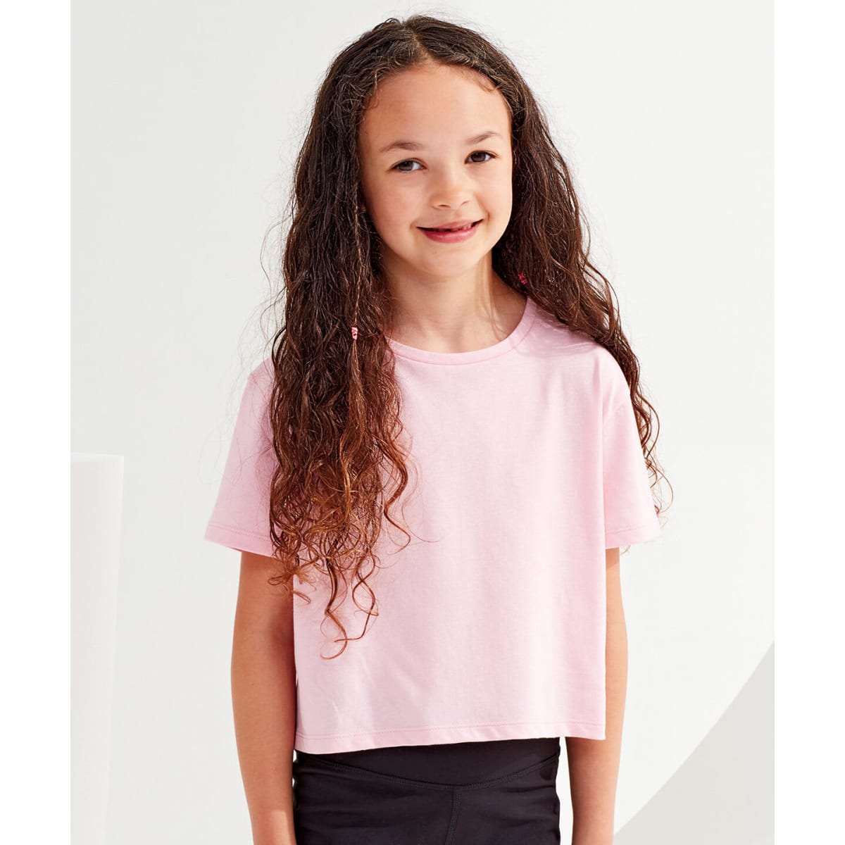 A pink kids boxy T-shirt on a girl with long, curly hair, standing against a light background.