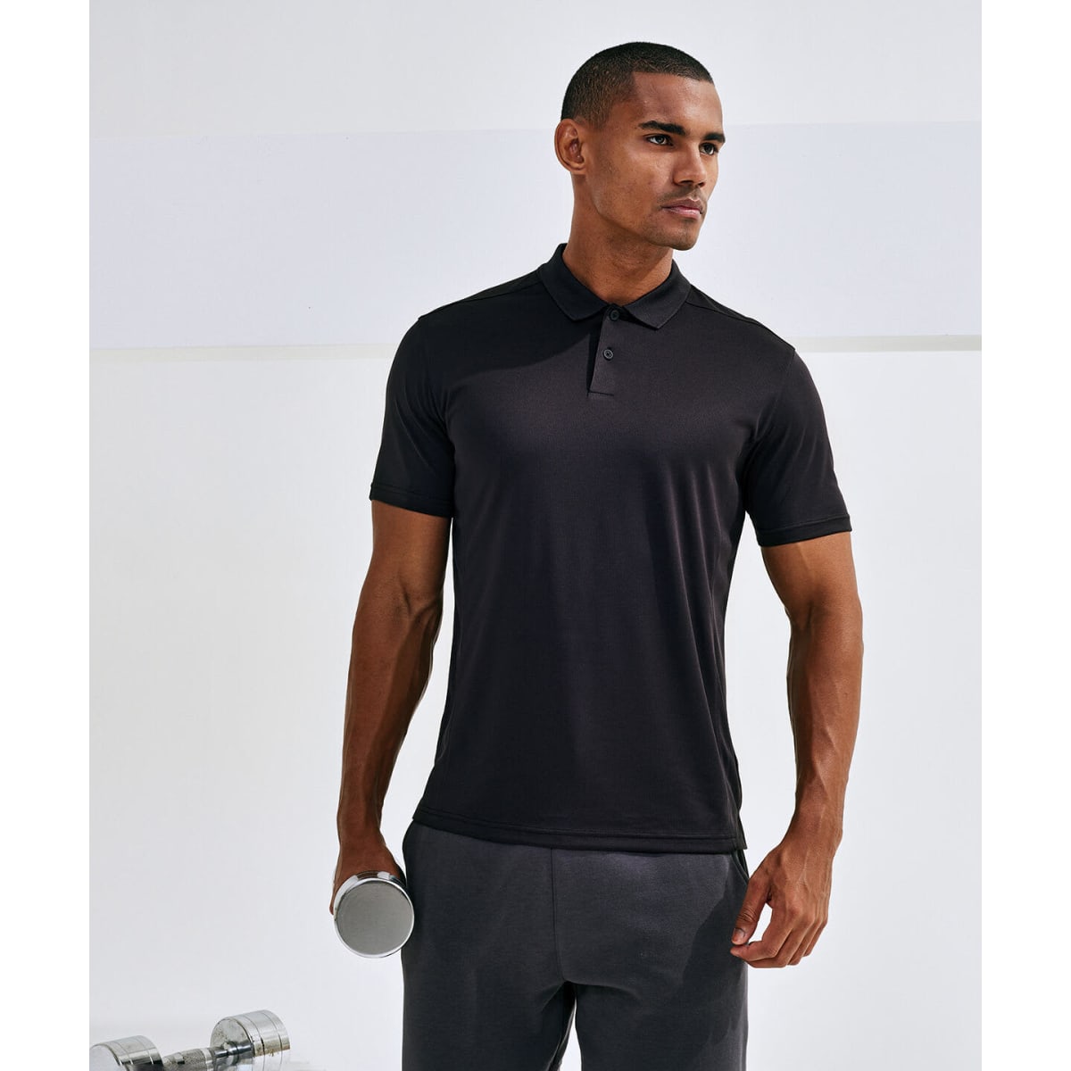 A man models a black Cationic Recycled Textured Polo, holding a dumbbell and wearing grey shorts. The image features a logo.