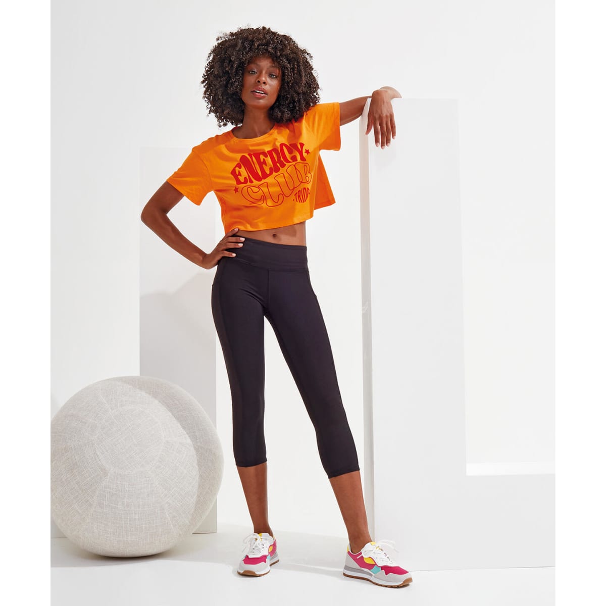 Black 3/4 length leggings displayed on a model, paired with an orange cropped t-shirt and colourful sneakers.