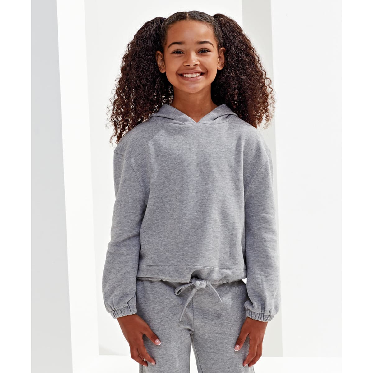 A grey, cropped oversize hoodie displayed on a smiling girl with curly hair, featuring a drawstring at the waist and a logo.