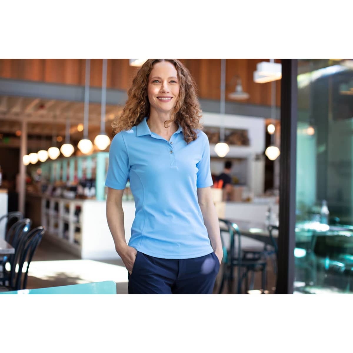 A woman stands in a café wearing a light blue polo shirt paired with dark trousers. The shirt features a logo.