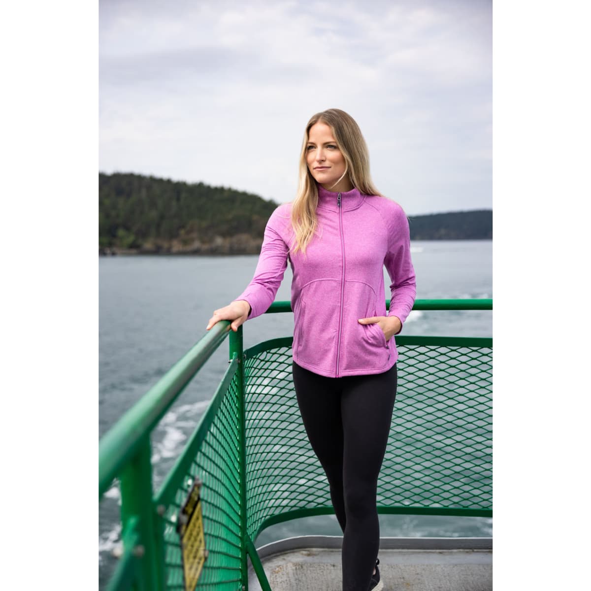 A woman stands on a boat wearing a pink full zip jacket and black leggings, with water and trees in the background.