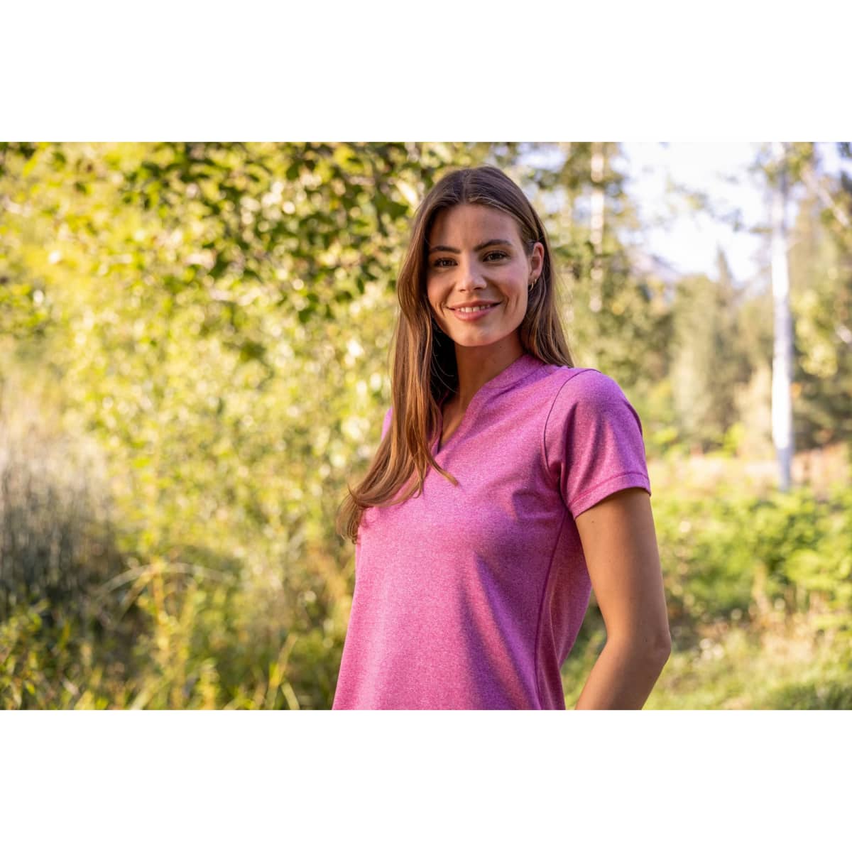A woman is wearing a pink polo shirt in a natural outdoor setting with trees in the background.