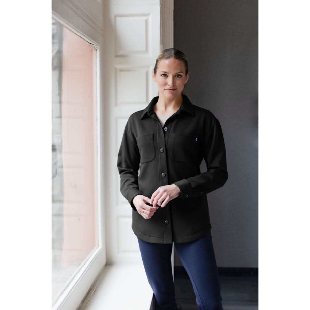 A black eco shirt jacket with a button-up front and two chest pockets, worn by a model indoors.