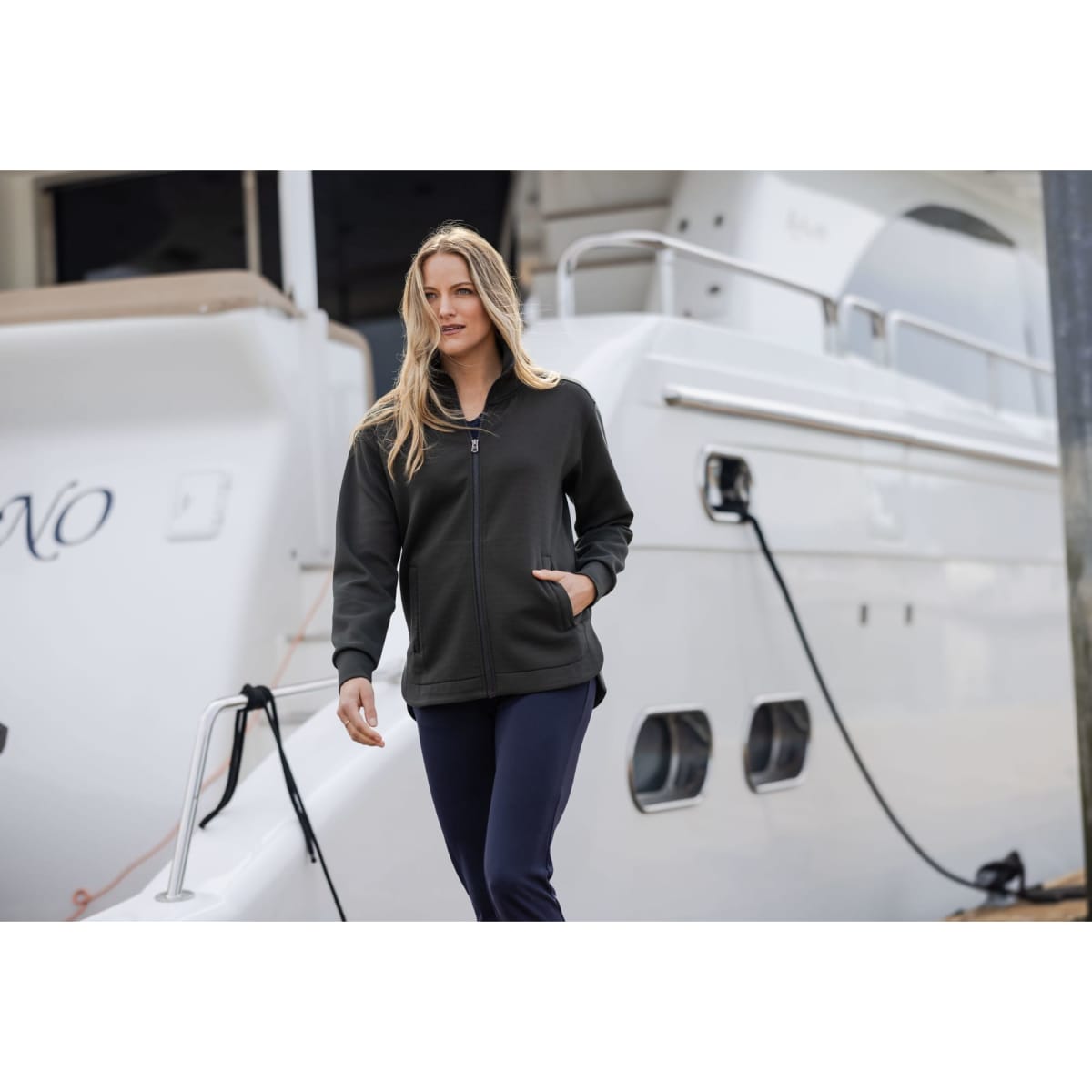 A woman in a dark grey full zip jacket walks by a yacht, showcasing the jacket's design and fit.