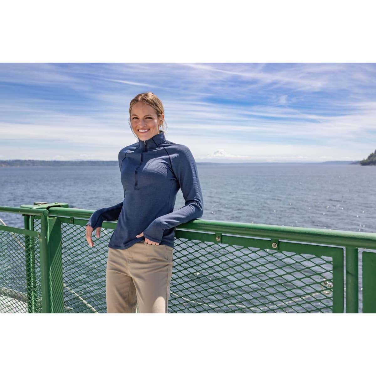 A woman stands on a ferry, wearing a blue eco fleece half zip jumper, with a scenic water view in the background.