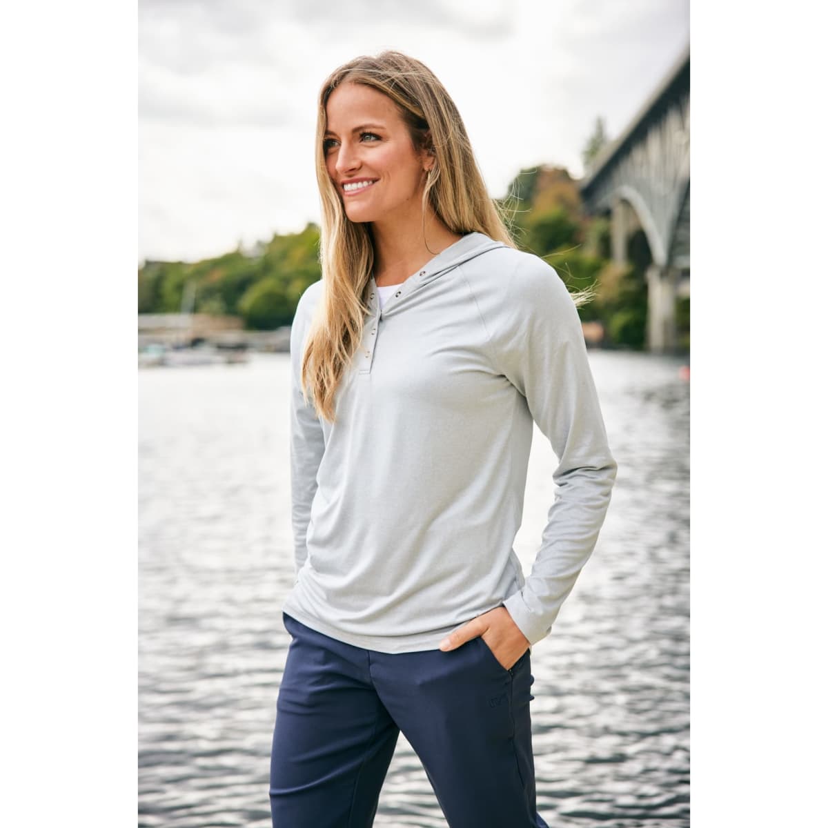 A hooded shirt in light grey, featuring long sleeves and a relaxed fit, displayed by a model near water.