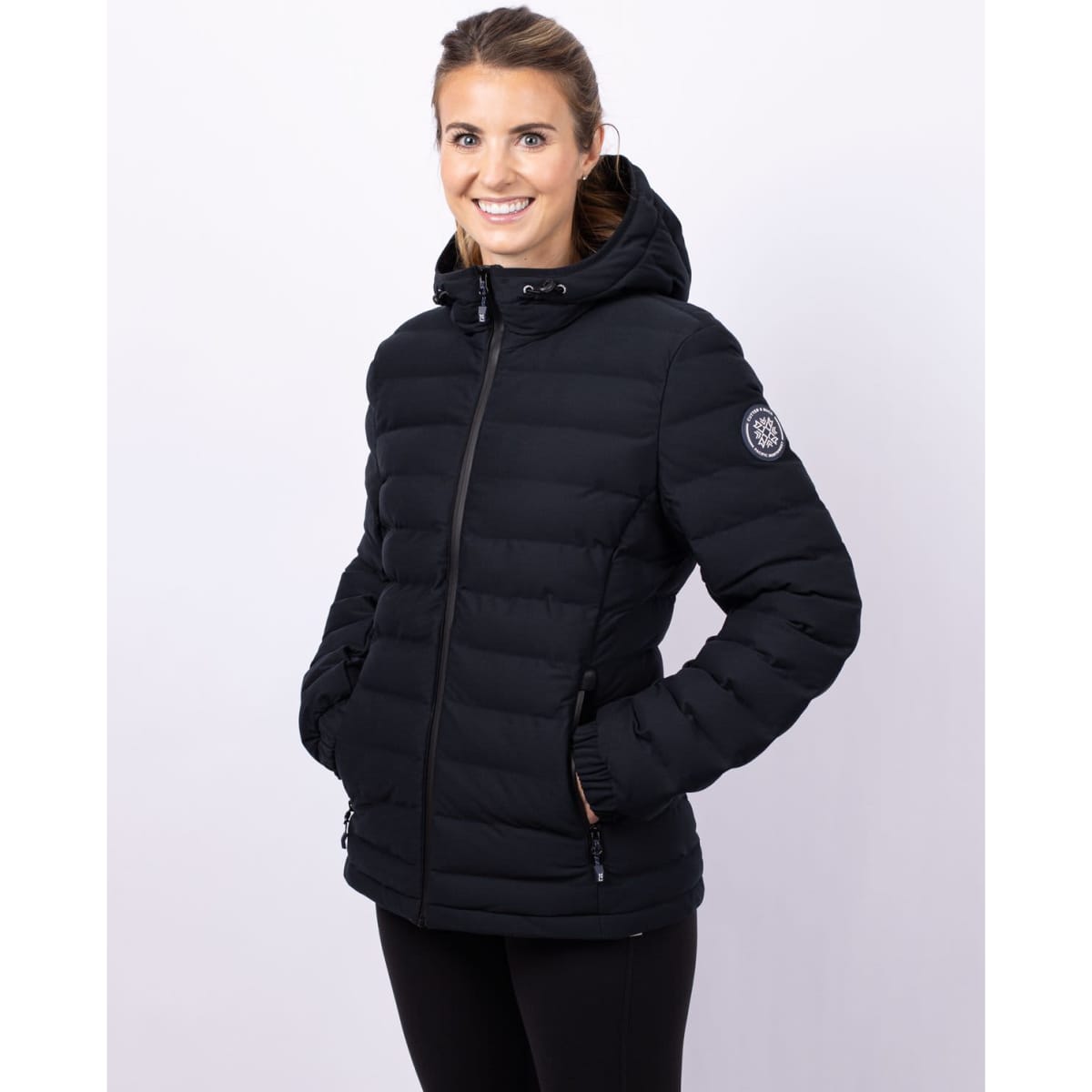 A black puffer jacket with a hood, featuring a logo on one sleeve, worn by a smiling woman.