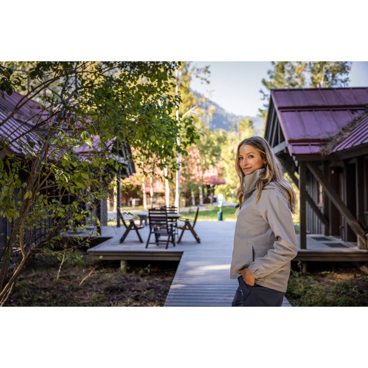 A light grey ladies full zip jacket is worn by a woman standing outdoors, surrounded by cabins and greenery.