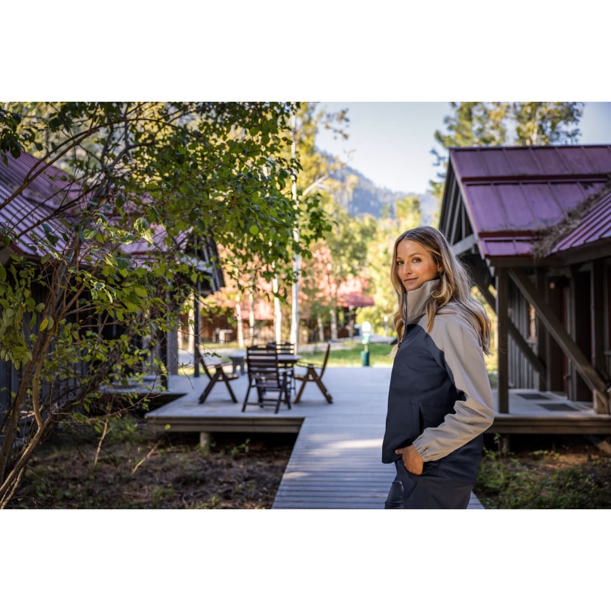A woman wearing a grey and navy rain jacket stands on a wooden path surrounded by cabins and trees.