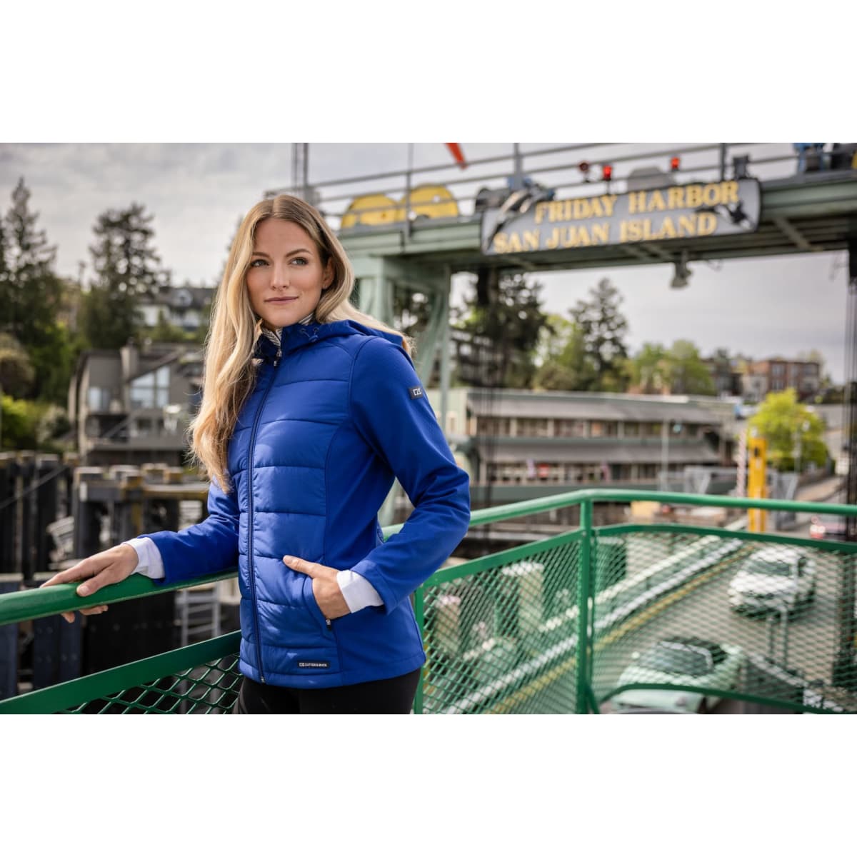 A woman wearing a blue soft shell jacket stands casually on a green railing with a scenic background.