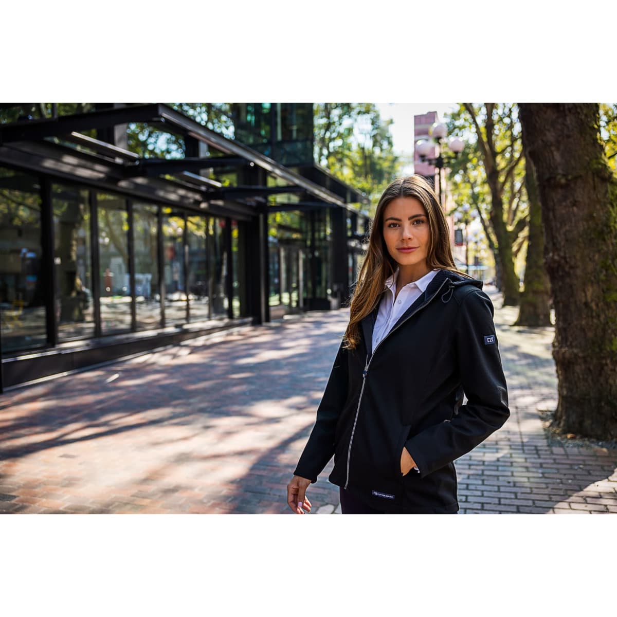 A woman stands outside wearing a black softshell jacket. The jacket features a hood and has a logo.