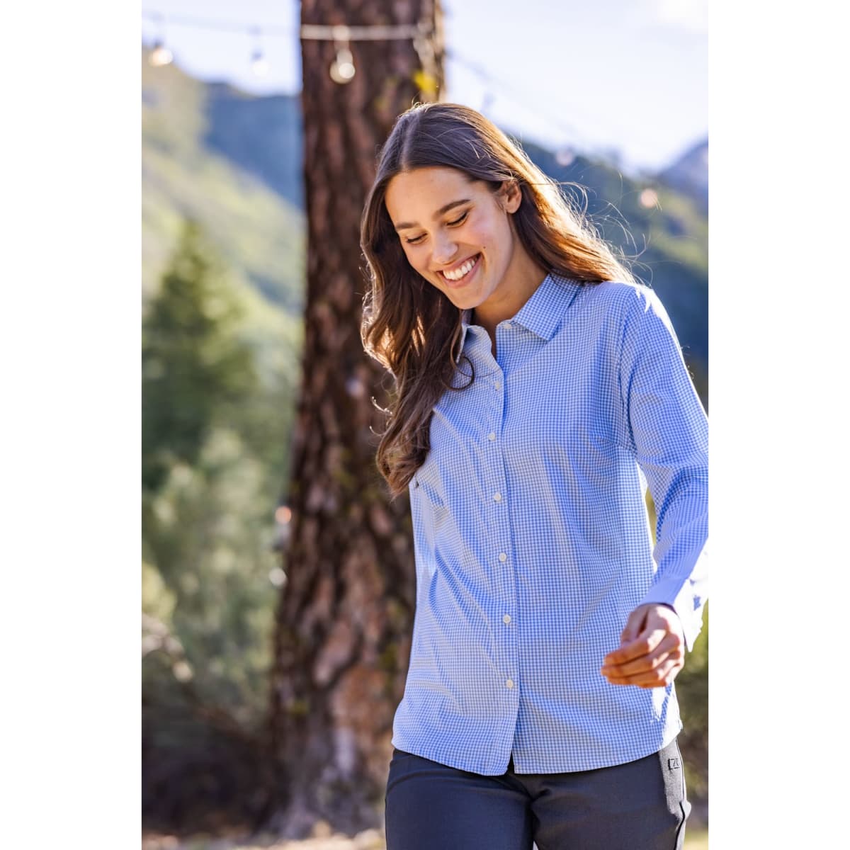 A woman is wearing a blue and white gingham shirt, smiling outdoors with trees in the background.